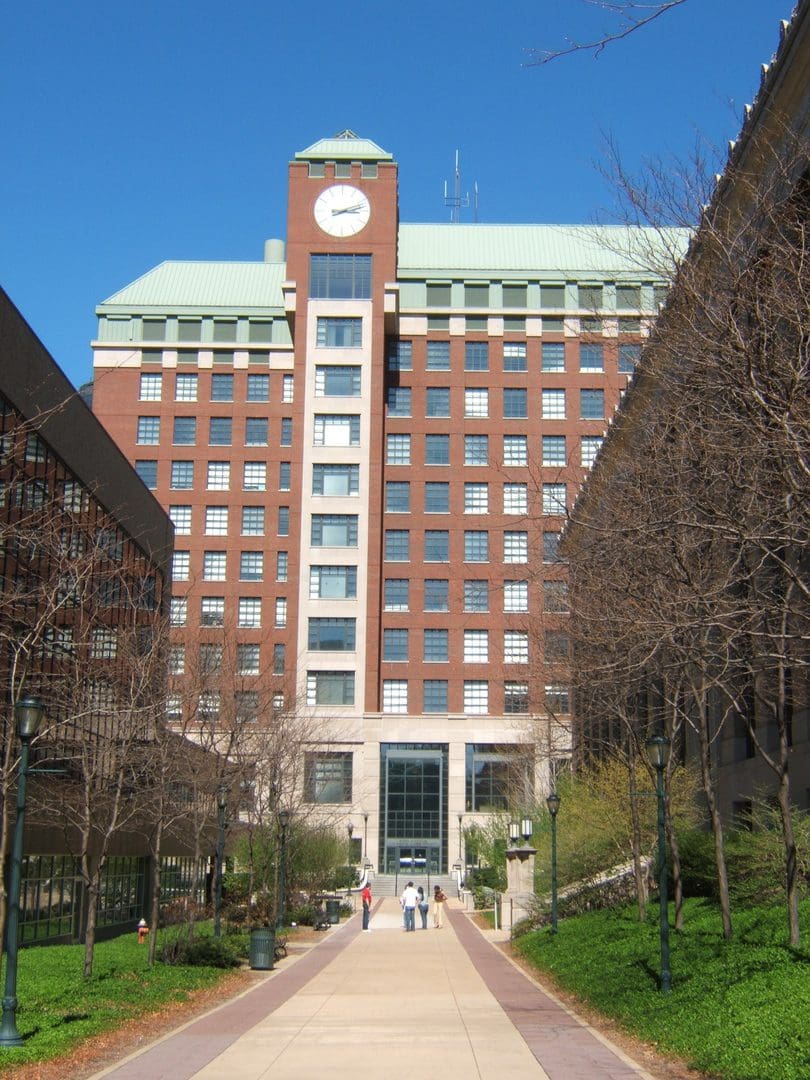 A tall brick building with a clock tower and trees lining a walkway.